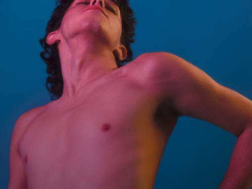 A shirtless person doing yoga with closed eyes, bathed in pinkish light against a blue background.