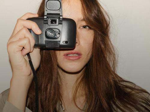 A woman with brown hair using an old camera in front of a grey wall, wearing an olive shirt.